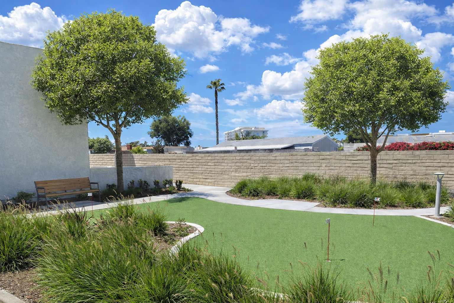 A small artificial putting green is surrounded by grass, shrubs, two trees, a bench, and a stone wall under a sunny, partly cloudy sky. - Four Seasons Landscaping