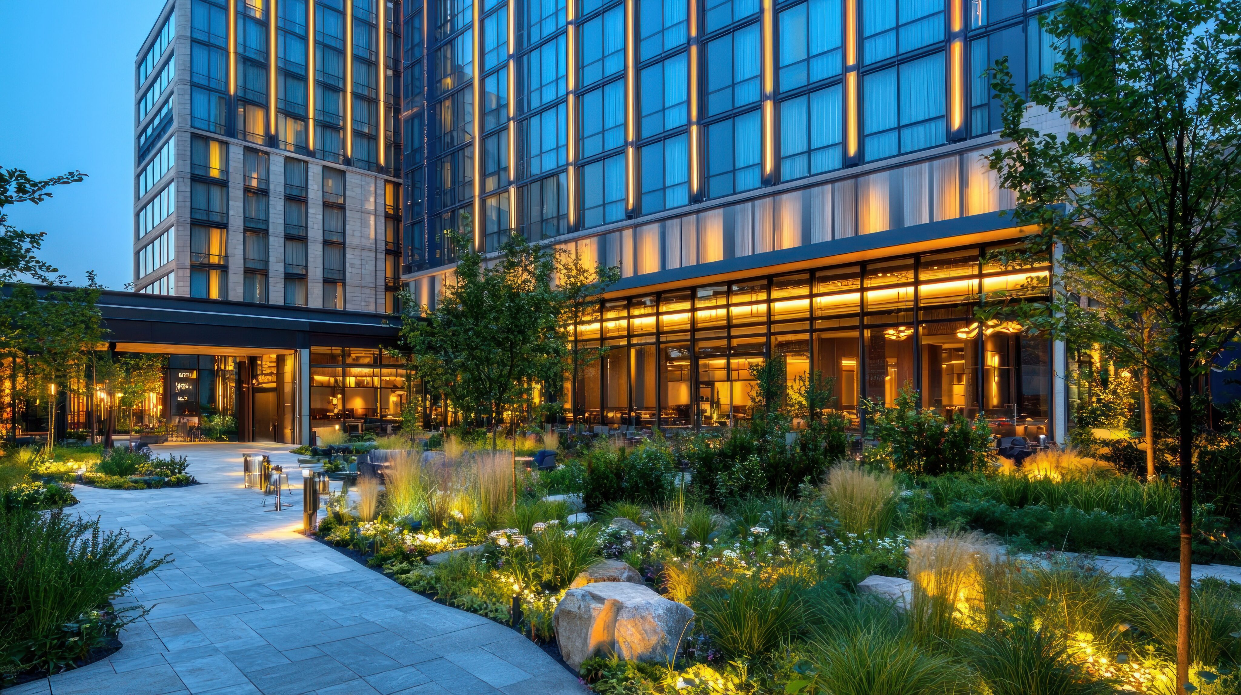 Modern hotel exterior at dusk with large windows, warm indoor lighting, and a landscaped garden featuring pathways, plants, and outdoor seating. - Four Seasons Landscaping